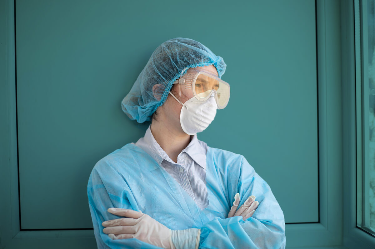 Photograph of a tired medical specialist with protective eyewear and PPF2 mask, leaning against a hospital wall with her arms folded while staring out a window