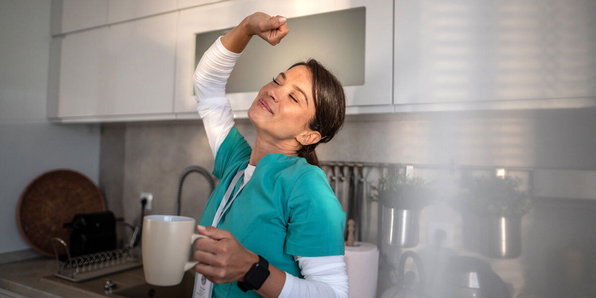 A young healthcare professional holding a hot drink and stretching whilst smiling in a home kitchen environment as early morning sunlight shines on the scene