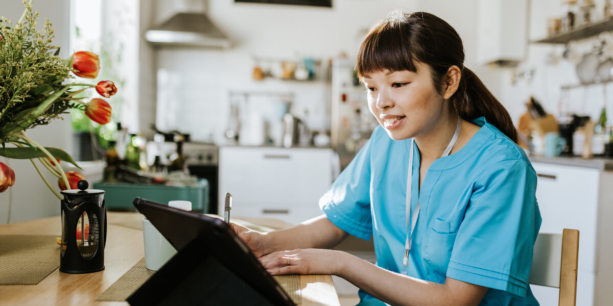 A young Asian healthcare professional writing whilst seated in a home kitchen environment and viewing a digital tablet device on the kitchen table